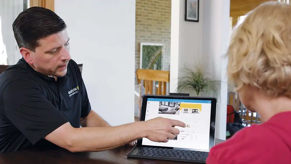 A man in a black Marvin Replacement polo shirt points to an image on a laptop with a customer. 