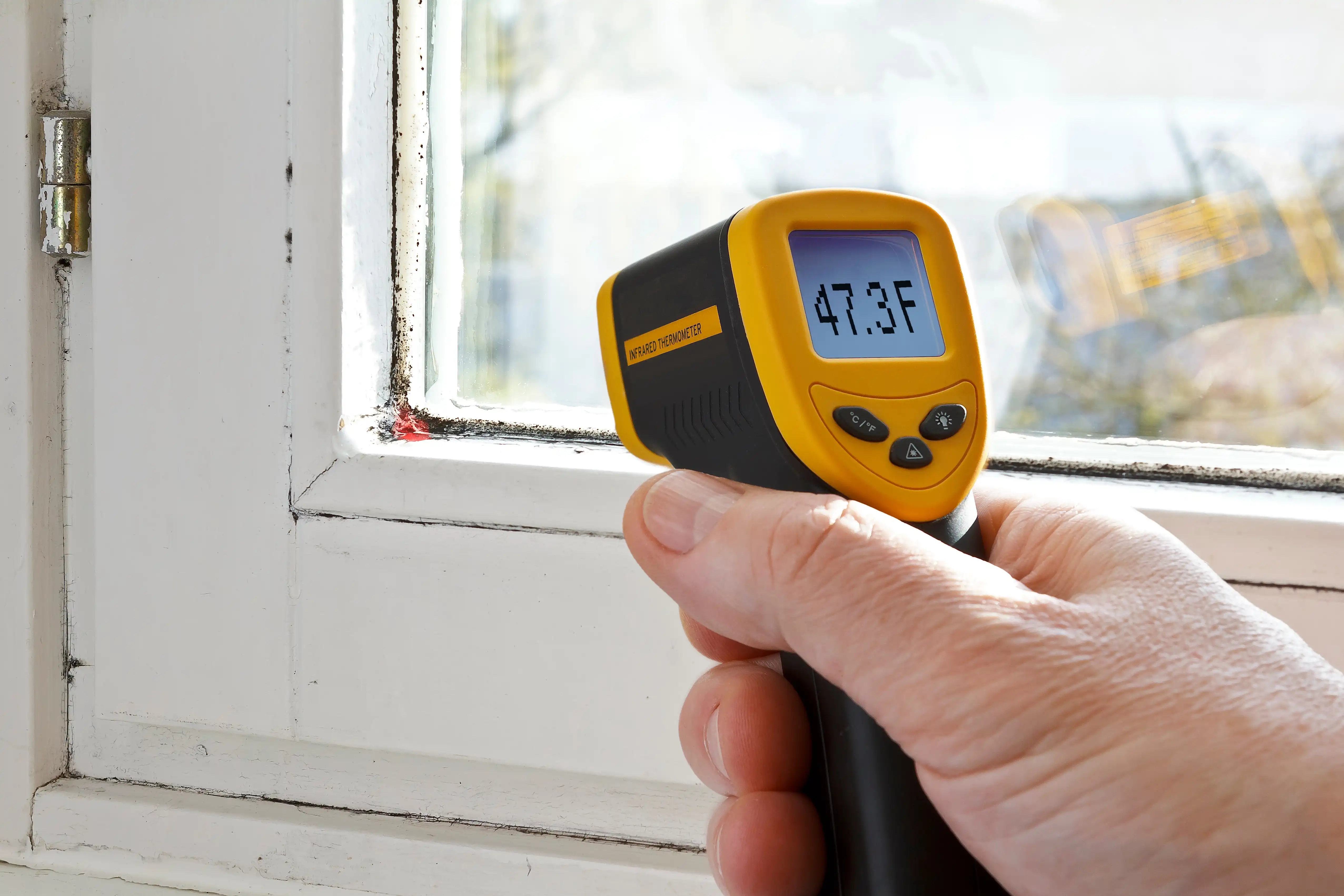 Image of a man using a thermal monitor to detect the temperate of the air near an old wood window.