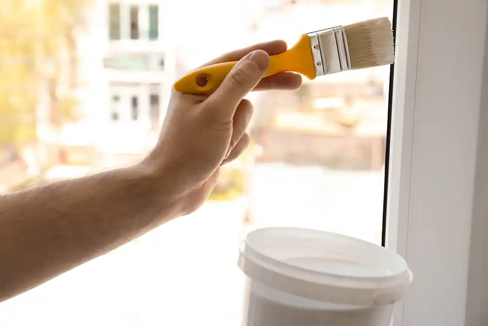 A man paints a window frame white with a yellow handled paint brush in his left hand.