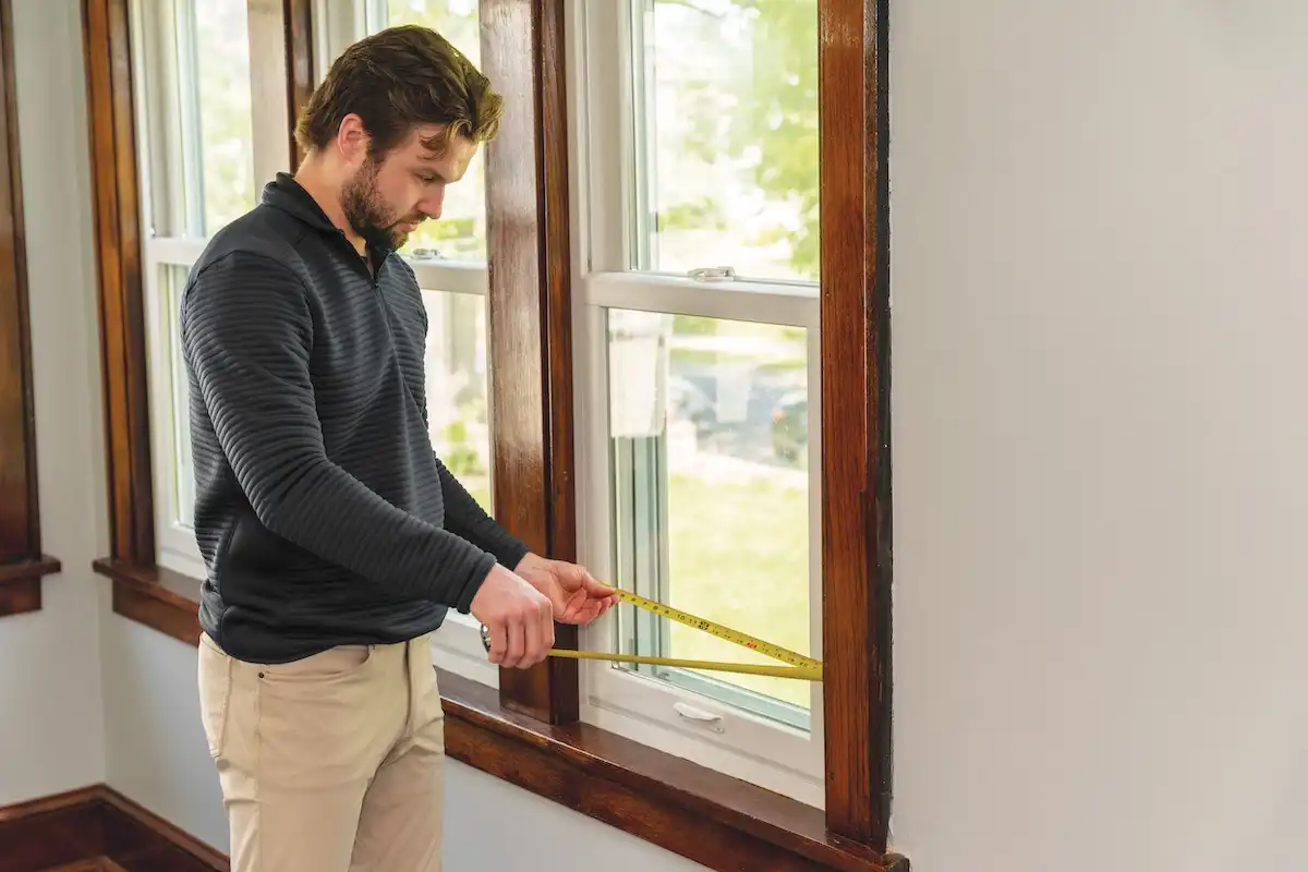 A man uses a tape measure to determine the width of a window.